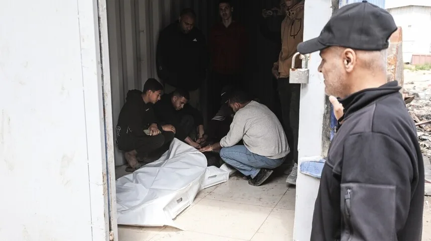 Grupa muškaraca okupljena oko tijela prekrivenog bijelom plahtom, unutar metalnog kontejnera.