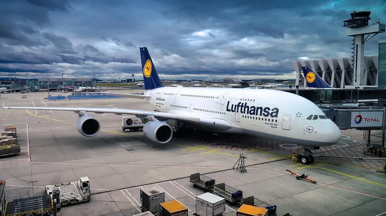 Lufthansa Airbus A380-800 na aerodromu pod tmurnim nebom, s vidljivim dijelovima terminala i tornjem za kontrolu leta.