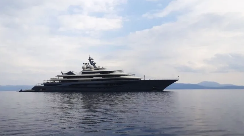 Velika luksuzna jahta tamne boje usidrena na mirnom moru, sa obrisima brda u pozadini pod oblačnim nebom.