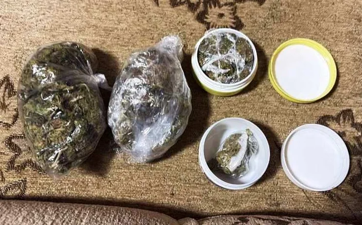 Dvije vrećice sa suhom marihuanom pored dvije otvorene bijele teglicama od kojih jedna sadrži marihuanu, a druga je prazna. Sve se nalazi na smeđoj tkanini sa uzorkom.