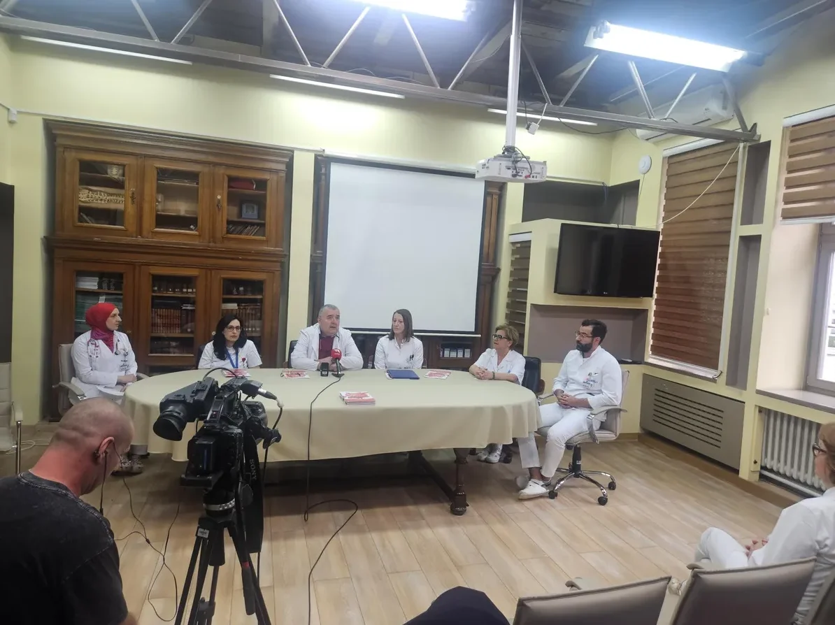 Grupa medicinskih radnika, uključujući nekoliko doktora u bijelim mantilima i jednu ženu u hidžabu, sjede za okruglim stolom na konferenciji ili sastanku. Ispred njih se nalaze mikrofoni, a u prvom planu stoji profesionalna kamera na stativu.