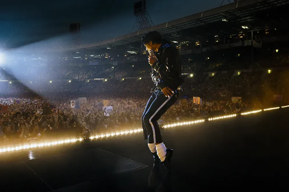 Michael Jackson u crnom odijelu sa bijelim čarapama i sjajnim pantalonama izvodi plesni pokret na osvijetljenom stage-u pred mnogobrojnom publikom u sportskoj dvorani.
