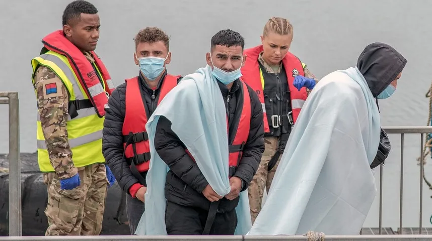 Grupa migranata, neki umotani u ćebad i noseći zaštitne maske, iskrcava se na obalu uz pomoć pripadnika oružanih snaga u prslucima za spašavanje.