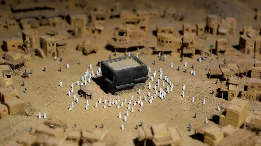 Minijaturni prikaz Kabe u Mekki okružene minijaturnim figurama ljudi u bijelim odorama, smještena u ambijentu sa minijaturnim zgradama od pustinjskog materijala.