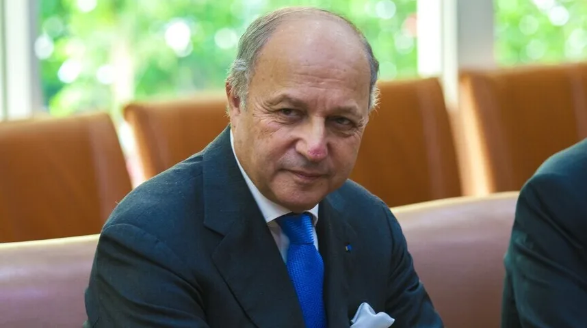 Portret uglednog gospodina u tamnom odijelu i plavoj kravati, kako sjedi za stolom sa drugim osobama u pozadini.