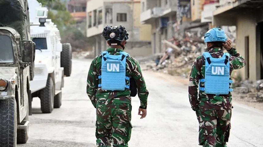 Mirovnjaci Ujedinjenih naroda (UN) u maskirnim uniformama i plavim prslucima s natpisom 'UN' hodaju ulicom pored niza vojnih vozila. U pozadini se vide uništene zgrade i ruševine, sugerišući područje pogođeno sukobom.