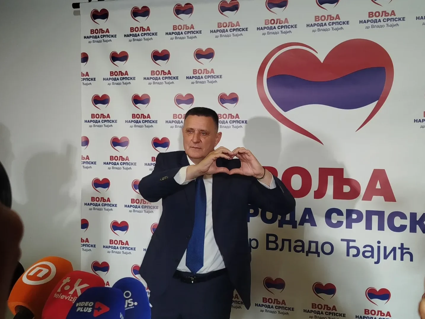 Мушкарац у тамном одијелу прави знак срца рукама испред позадине са натписом "Воља народа српске др Владо Бајић" и симболом срца у бојама заставе Србије.