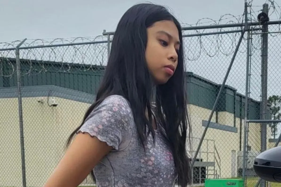 Mlada djevojka sa dugom crnom kosom nosi sivu majicu kratkih rukava sa cvjetnim dezenom, stojeći ispred ograde sa bodljikavom žicom.