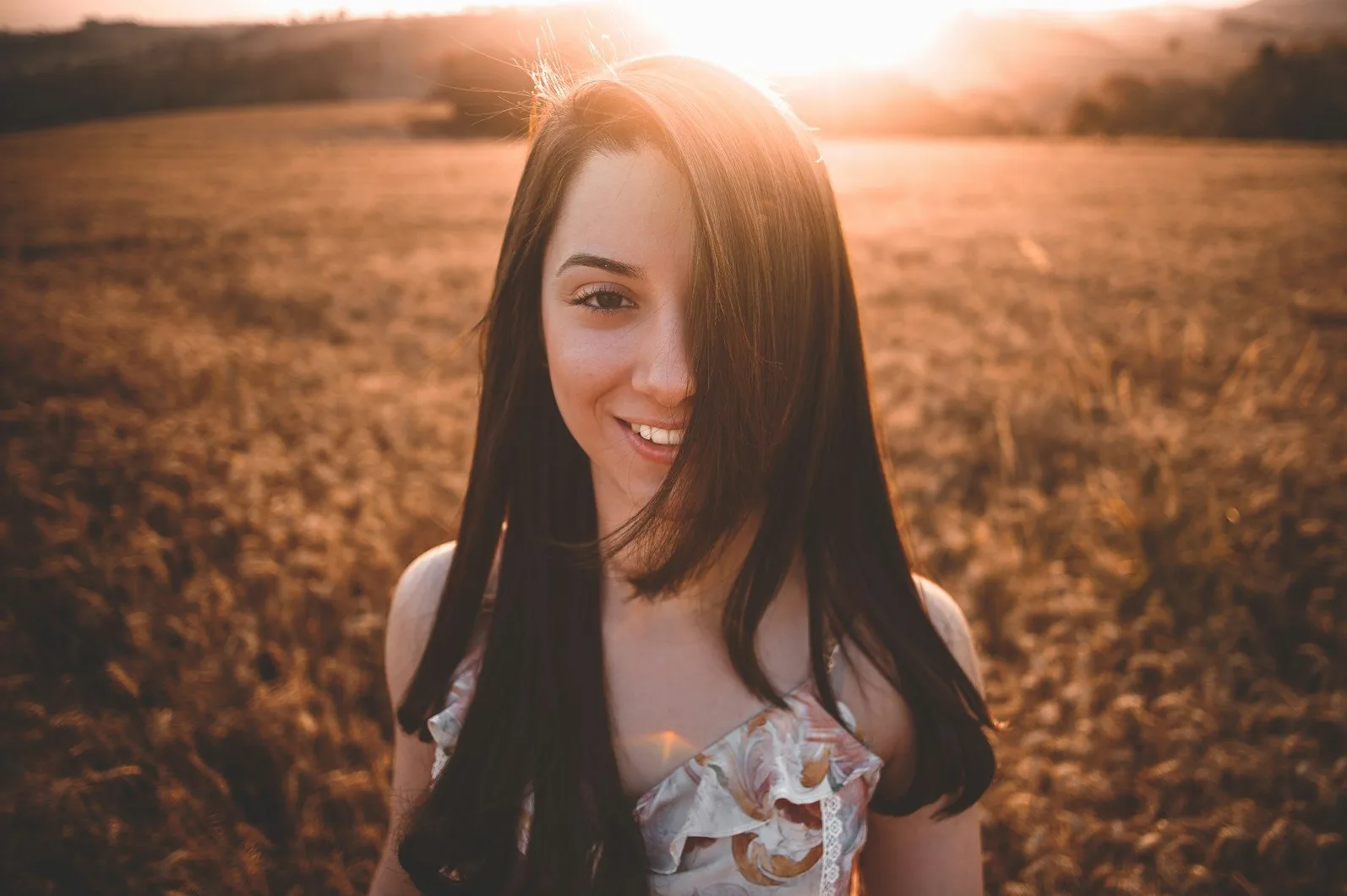 Portret mlade djevojke sa dugom tamnom kosom kako se smiješi u polju pšenice tokom zalaska sunca.
