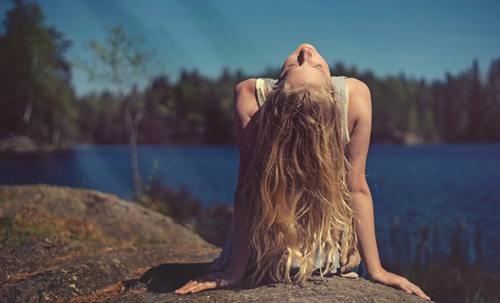 Mlada djevojka sa dugom plavom kosom naginje glavu unazad dok sjedi na stijeni pored jezera.