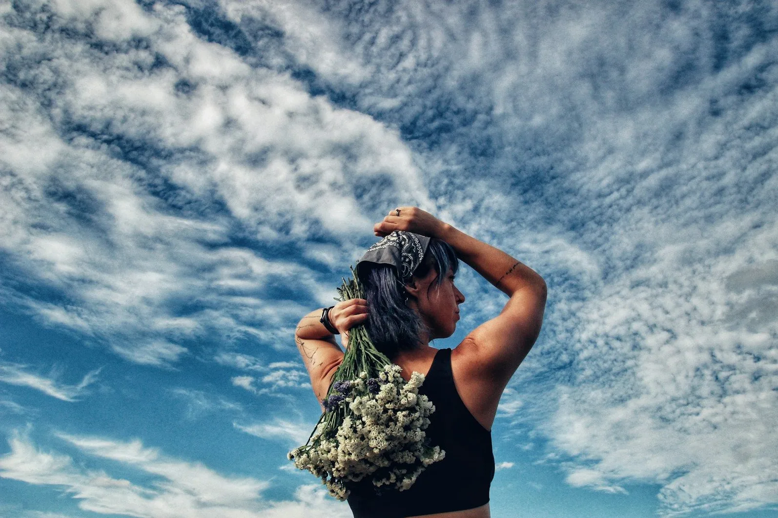 Žena sa ljubičastom kosom, crnom majicom i maramom na glavi, drži buket cvijeća iza leđa i gleda u nebo