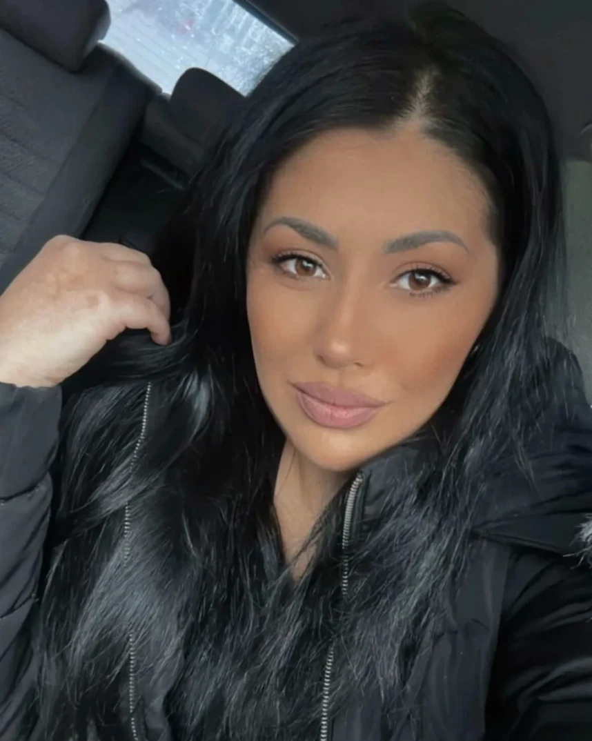 Portret mlade žene sa dugom crnom kosom, smeđim očima i ružičastim usnama, obučenoj u crnu jaknu, dok sjedi u automobilu.