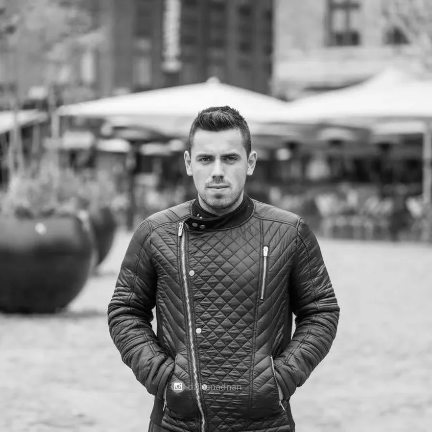 Crno-bijela fotografija mladog muškarca sa rukama u džepovima, obučenog u prošivenu jaknu.