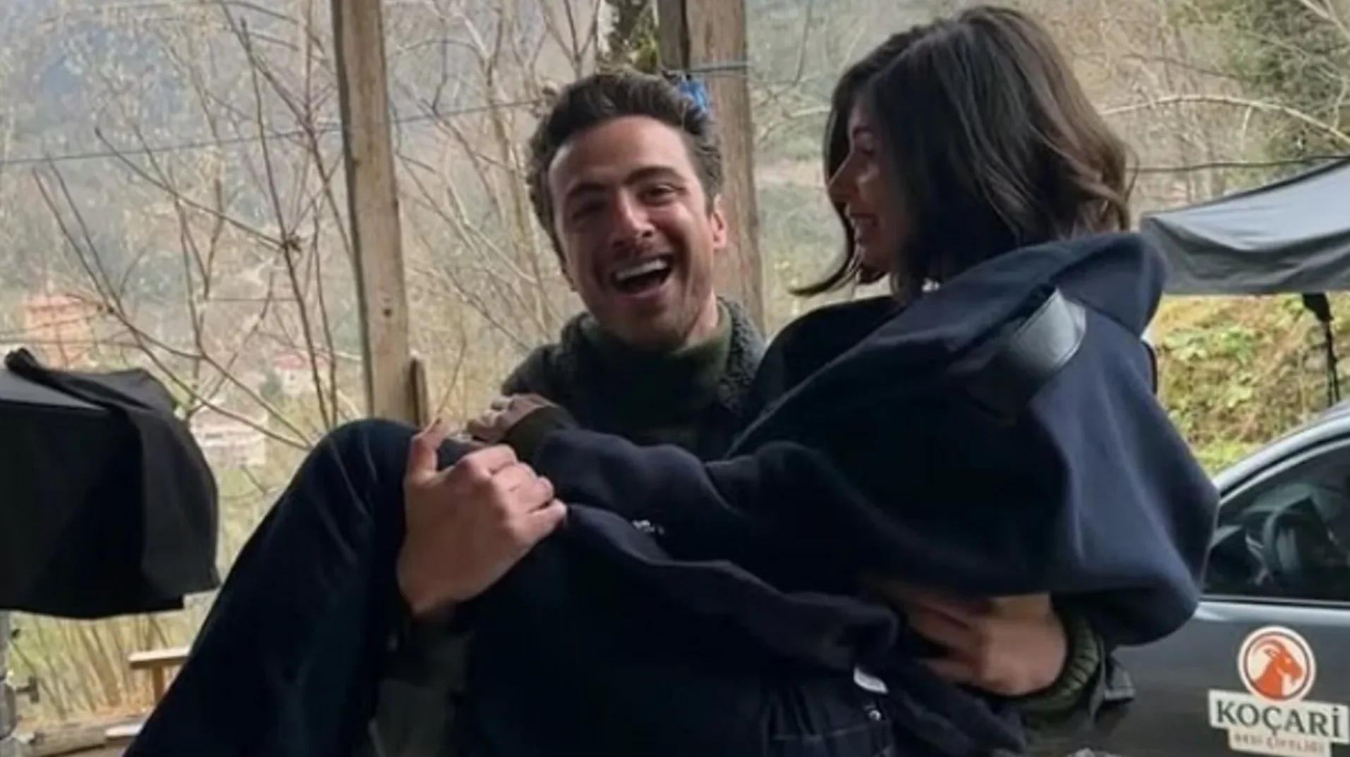 Mladić veselo drži djevojku u naručju na otvorenom, na lokaciji nalik snimanju.