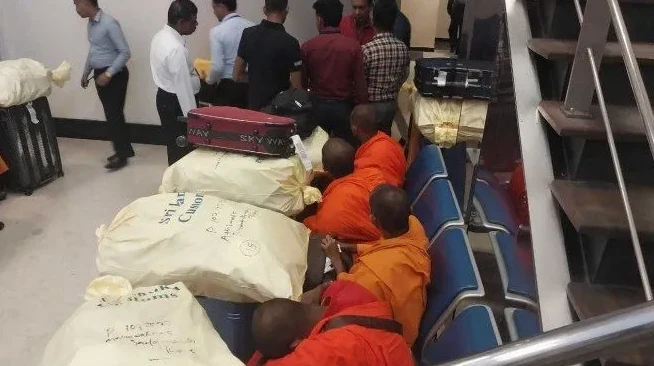 Grupa monaha u narandžastim haljinama sjedi pored obilne prtljage na aerodromu.