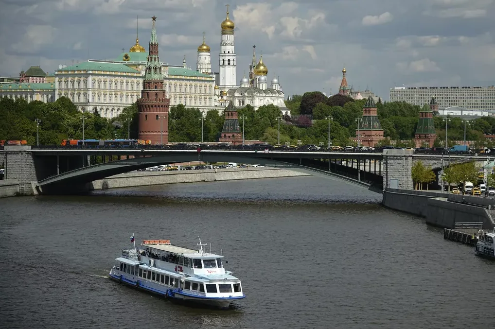 Pogled na Moskovski Kremlj s kupolama i tornjevima iznad rijeke s brodom u prvom planu i mostom punim automobila i autobusa.