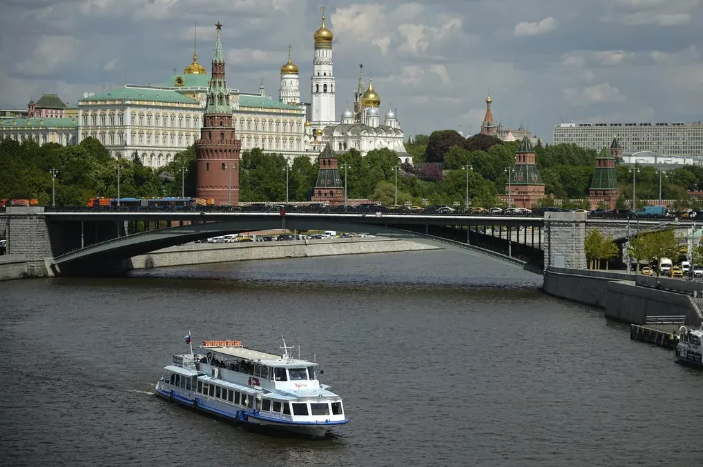 Pogled na Moskovski kremlj sa zlatnim kupolama i tornjevima, premošćen mostom preko rijeke Moskve, s brodom koji plovi rijekom.