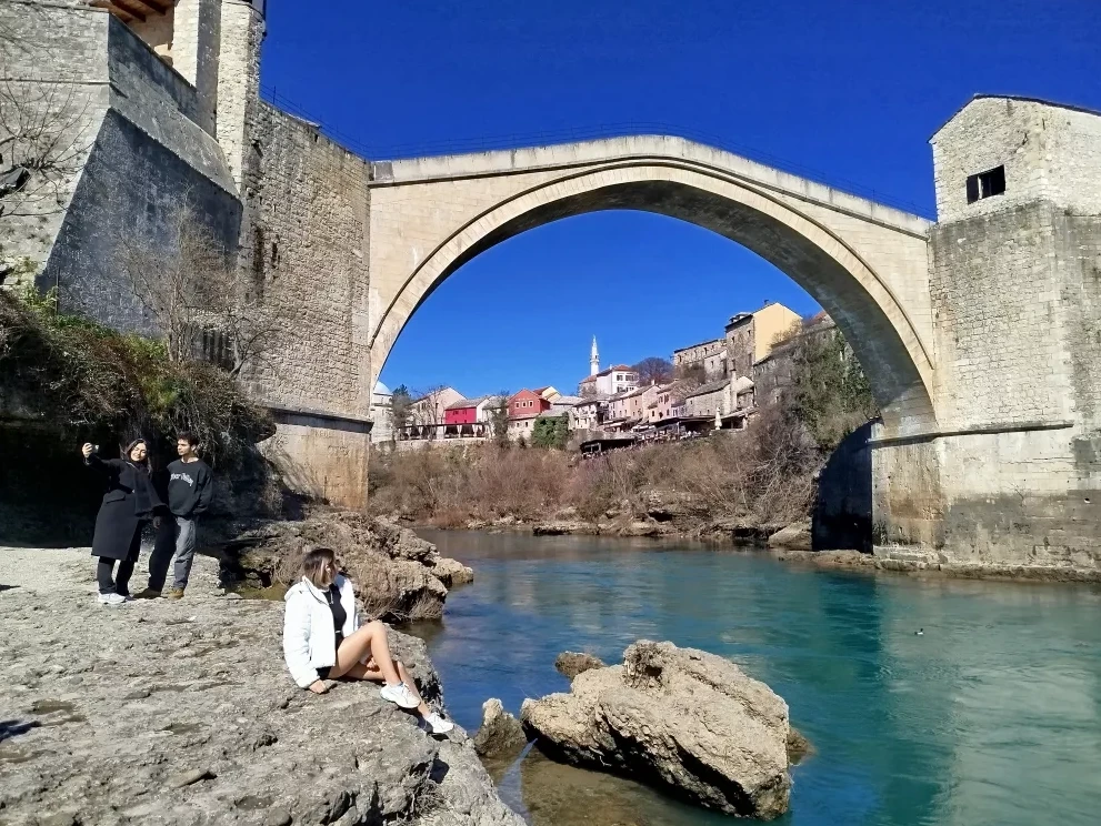 Tri osobe sjede i stoje na stijenama pored rijeke Neretve, sa Starim mostom u Mostaru, Bosna i Hercegovina, na sunčan dan.