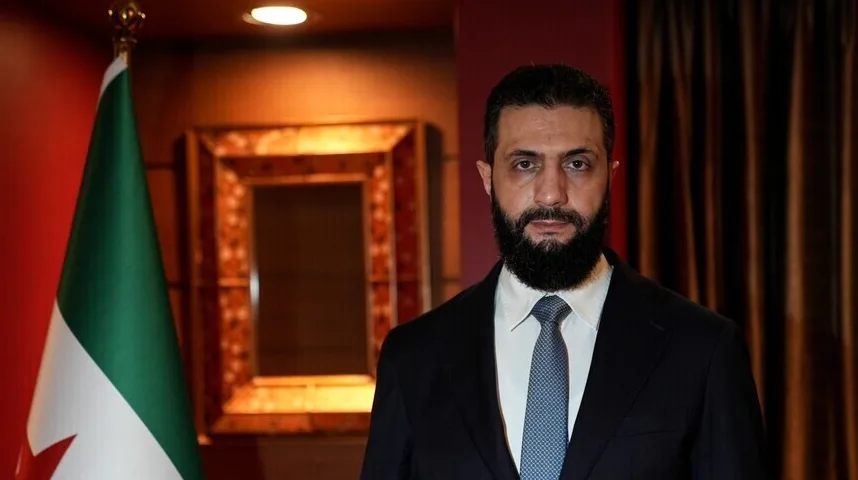 Muškarac sa bradom i tamnim odijelom, stojeći ispred zastave i ukrašenog okvira.