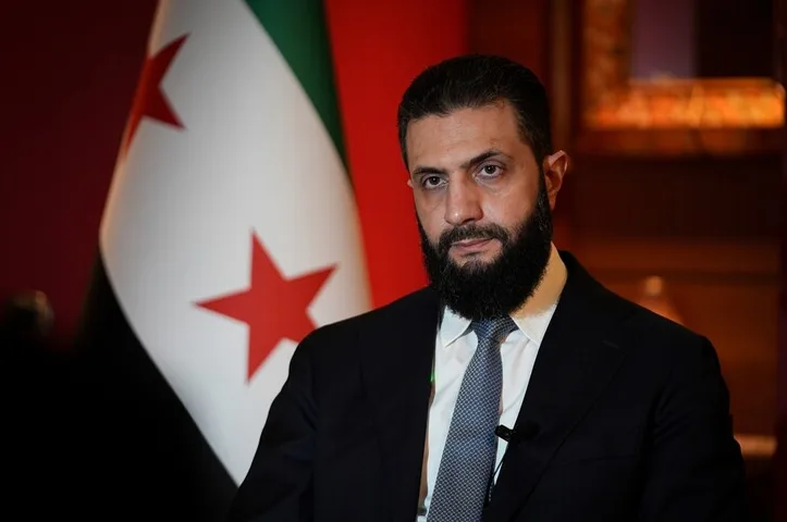 Muškarac sa bradom u crnom odijelu i plavoj kravati ispred zastave Sirije.