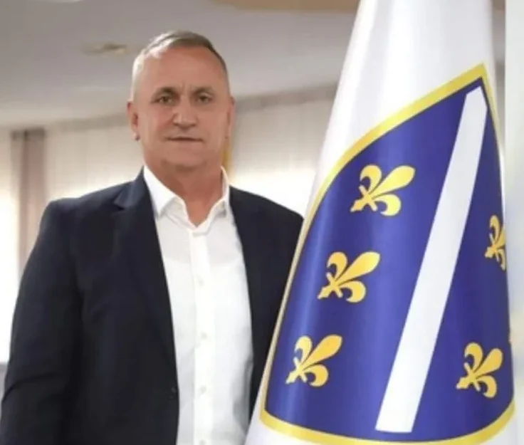 Portret muškarca u tamnom odijelu i bijeloj košulji, stojećeg pored zastave Bošnjaka.