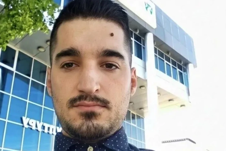 Mladić sa urednom frizurom, bradom i mladežom na čelu, nosi tamnoplavu majicu s bijelim tačkicama. Pozira ispred moderne zgrade sa staklenim fasadama i zelenilom u pozadini.