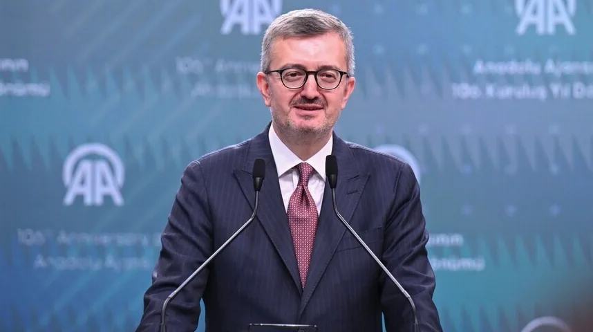 Muškarac sa naočarima i kravatom stoji iza podijuma sa dva mikrofona, na zelenoj pozadini sa logom "AA" i natpisom "Anadolu Ajansı 100. Kurulus Yilı Dra".