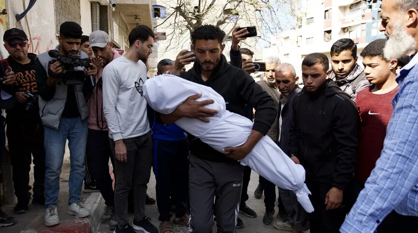 Muškarac u crnoj dukserici nosi umotano tijelo u bijeli pokrovac, okružen ljudima koji gledaju i snimaju kamerama i mobilnim telefonima.