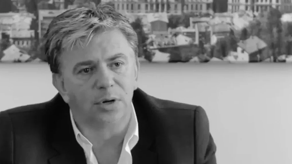 Portret srednjovječnog muškarca u crno-bijeloj tehnici, obučenog u tamno odijelo i bijelu košulju, kako govori.