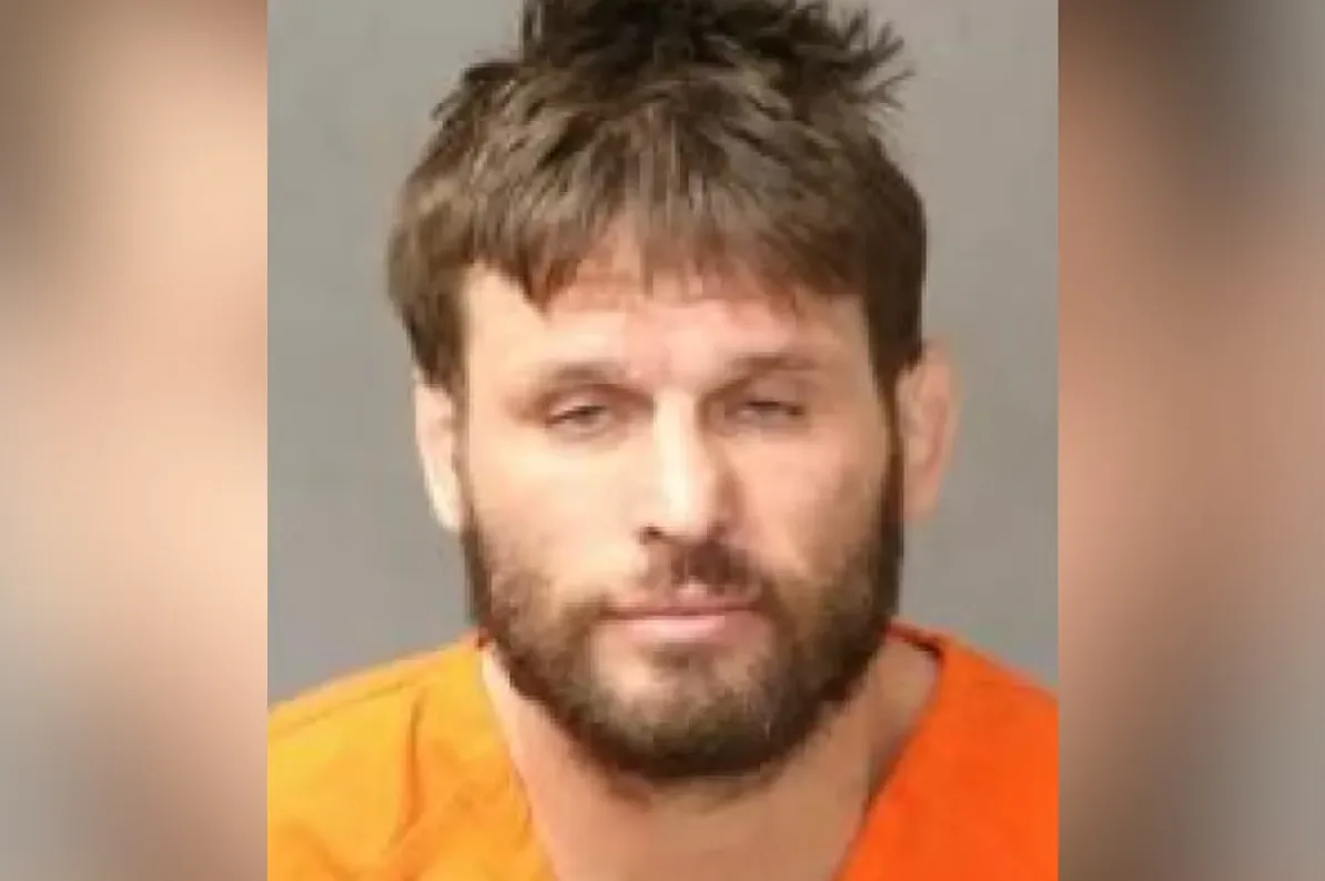 Portret muskarca sa kratkom smedjom kosom i bradom, obučenog u narandžastu zatvorsku odjeću.