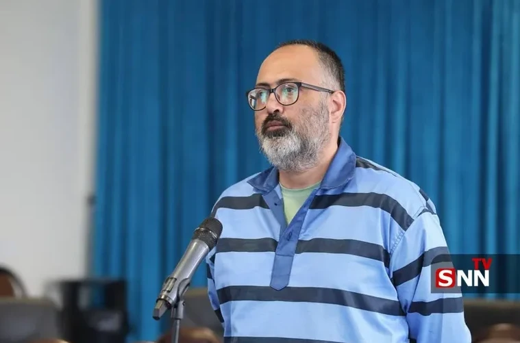 Muškarac sa naočalama i bradom, obučen u plavo-crnu prugastu košulju, stoji ispred mikrofona i govori.