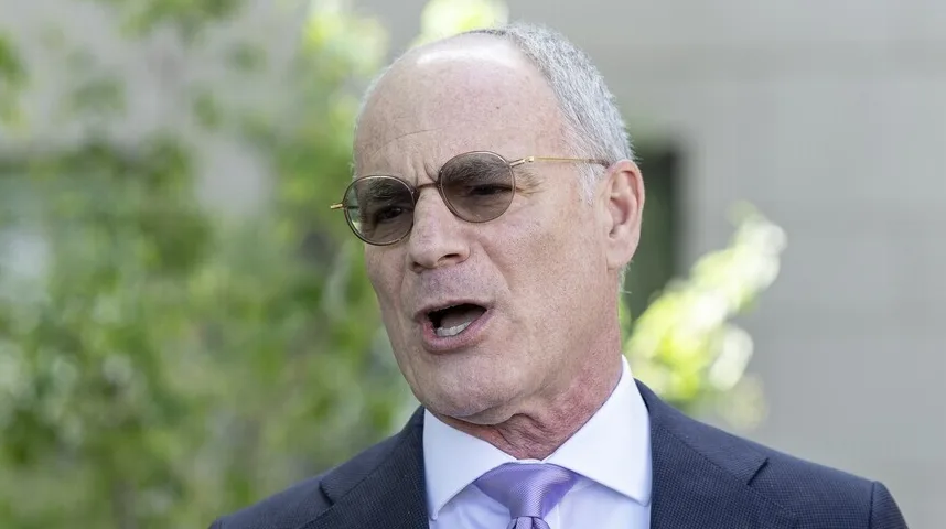 Portret starijeg muškarca s kratkom sijedom kosom, okruglim smeđim naočalama i ljubičastom kravatom, koji govori napolju.