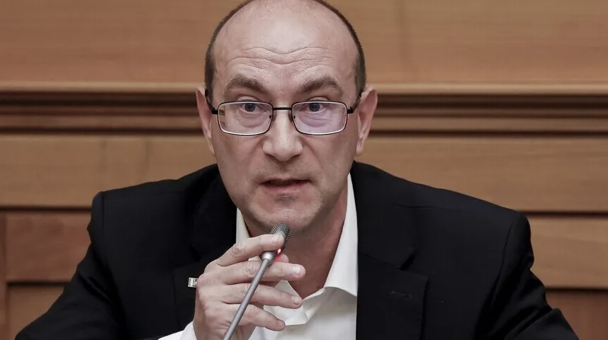 Portret muškarca s naočarima i crnim odijelom, koji govori u mikrofon.