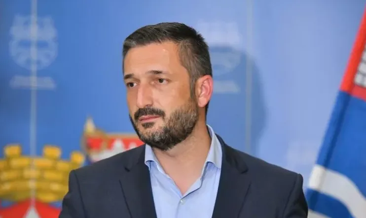 Portret muškarca sa kratkom tamnom kosom, urednom bradom, obučenog u tamno odijelo i svijetloplavu košulju. Pozadina je plava sa elementima grba i zastave.