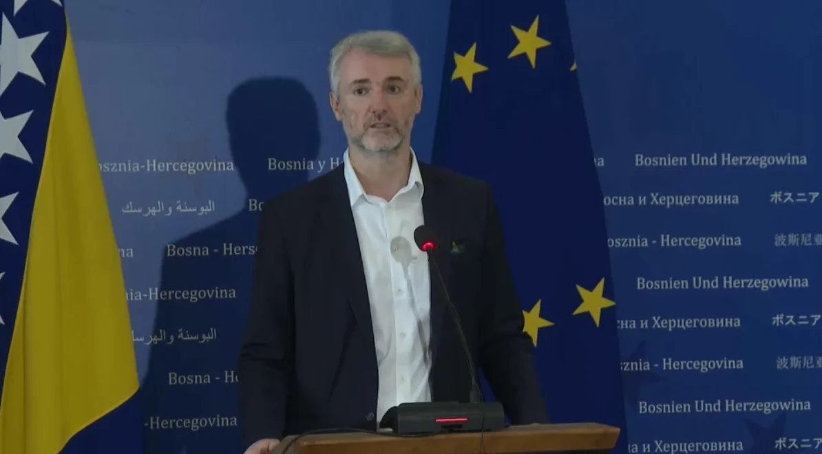 Čovjek sa sijedom kosom i bradom, obučen u tamno odijelo i bijelu košulju, govori za govornicom.