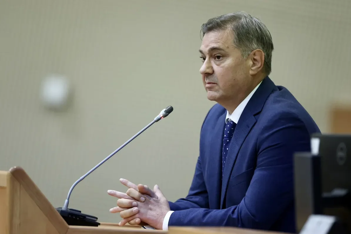 Čovjek u tamnoplavom odijelu sa bijelom košuljom i plavom kravatom sa tačkicama, sjedi iza drvenog podijuma sa mikrofonom ispred sebe.