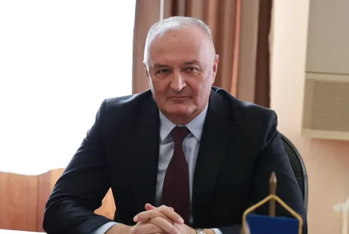 Portret starijeg muškarca u tamnom odijelu i tamnoj kravati, sjedi za stolom sa rukama prekriženim.