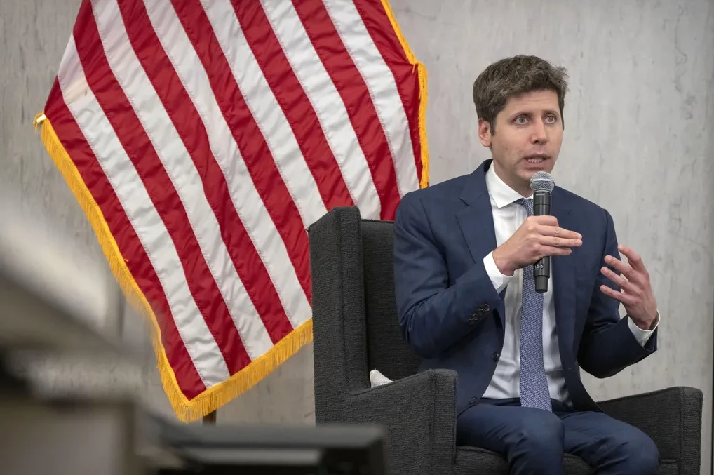 Muškarac u tamnom odijelu i bijeloj košulji sa plavom kravatom sjedi i drži mikrofon u desnoj ruci dok gestikulira lijevom rukom. U pozadini je zastava Sjedinjenih Američkih Država.