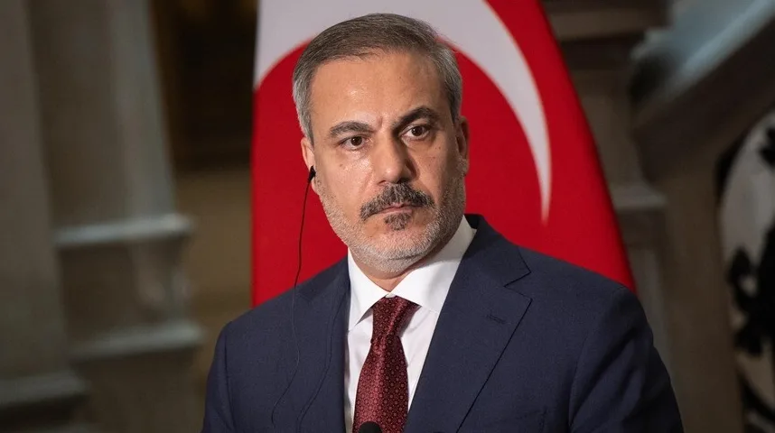 Muškarac u tamnom odijelu i crvenoj kravati, sa slušalicom u uhu, stoji ispred turske zastave.