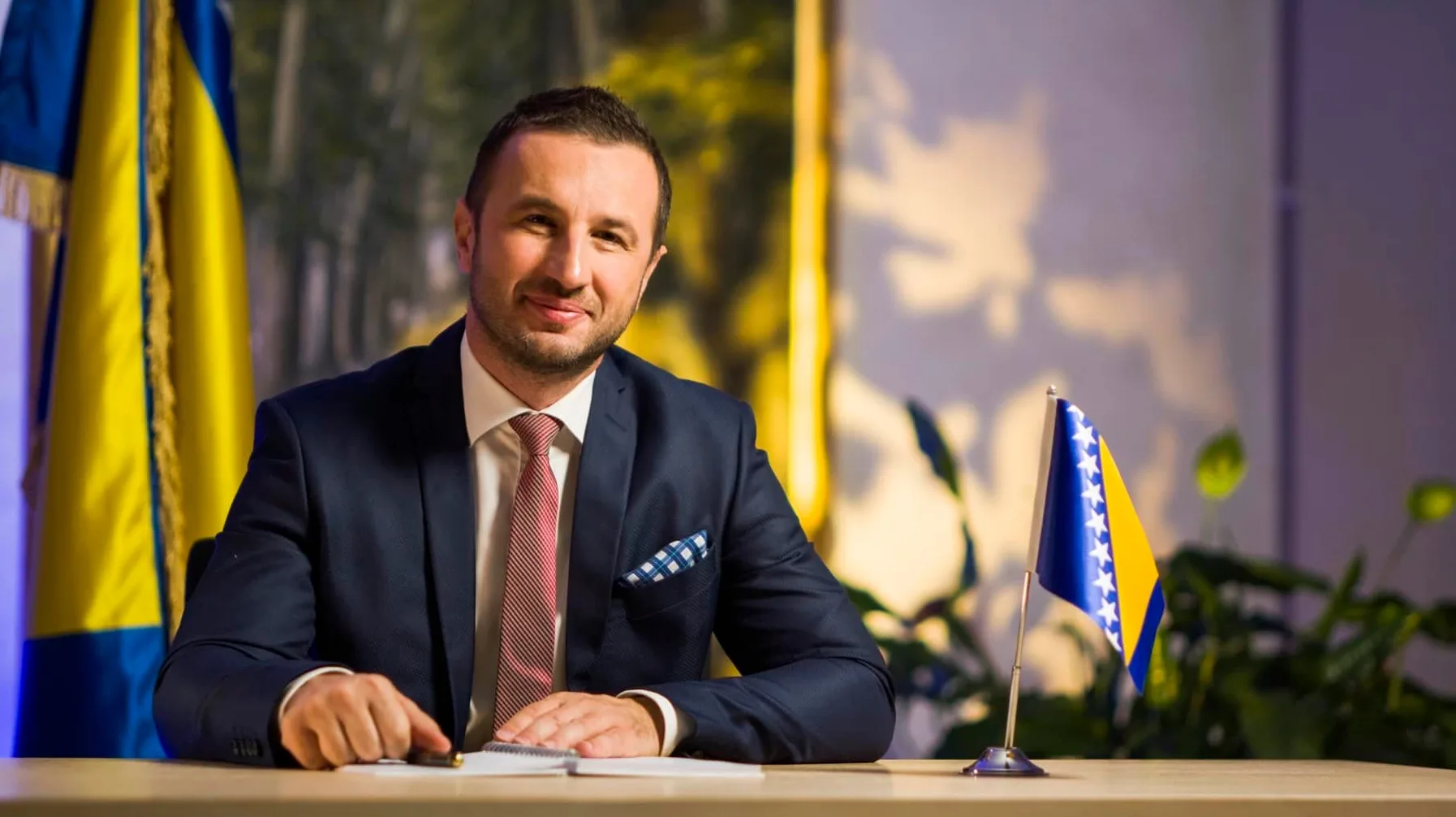 Političar ili službenik u tamnom odijelu, bijeloj košulji i crvenoj kravati, sjedi za stolom ispred zastave Bosne i Hercegovine.