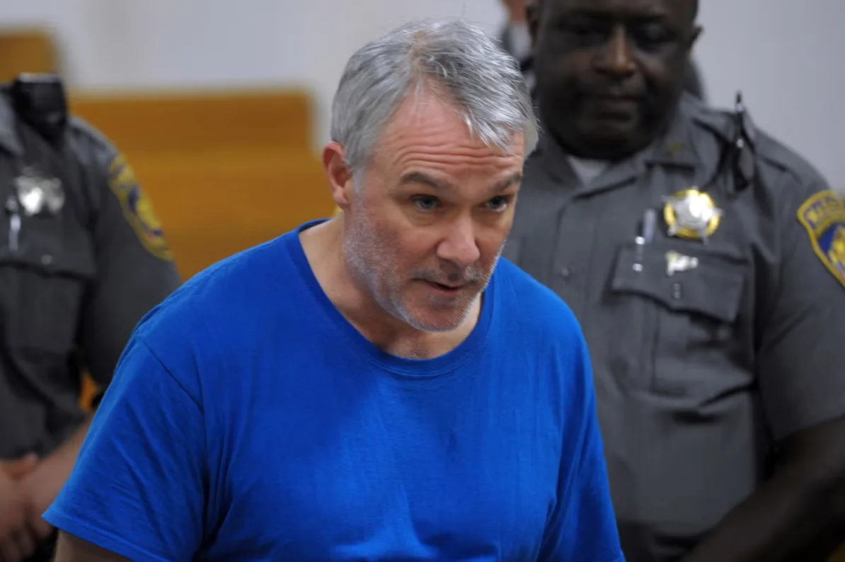 Muškarac sa sijedom kosom i bradom u plavoj majici, okružen policajcima u uniformama.