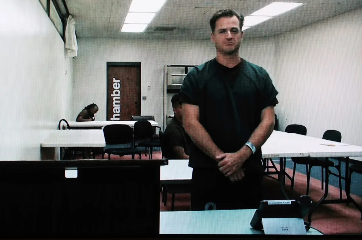 Muškarac u zatvorskoj uniformi stoji pred kamerom u prostoriji za videokonferencije, vjerovatno na suđenju.