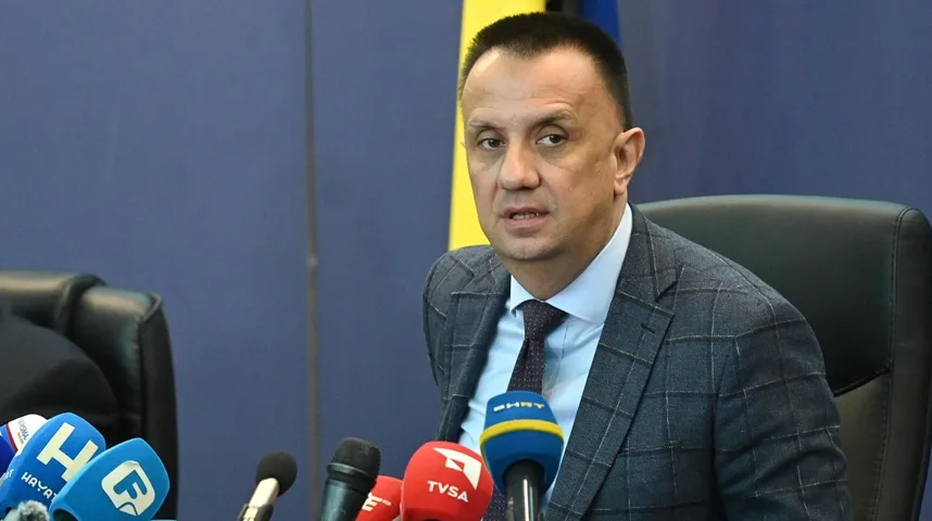 Portret političara koji sjedi za stolom ispred nekoliko mikrofona medija.