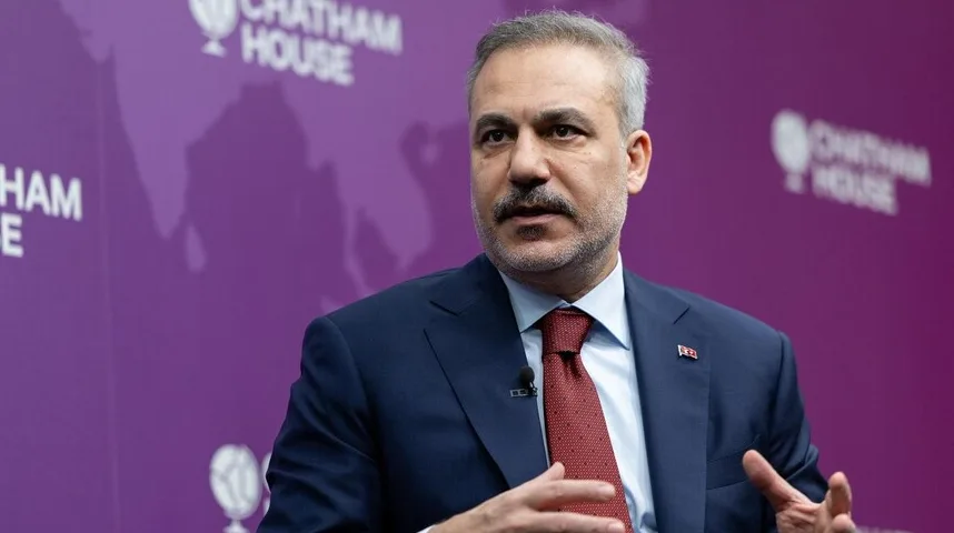 Čovjek u tamnoplavom odijelu i crvenoj kravati sa uzorkom, sa sijedom kosom i brkovima, gestikulira rukama dok govori. U pozadini se jasno vidi ljubičasta boja sa logom "Chatham House" ispisanim bijelim slovima.