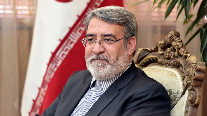 Portret poznate osobe s bradom i naočalama, sjedi u fotelji pored iranske zastave.