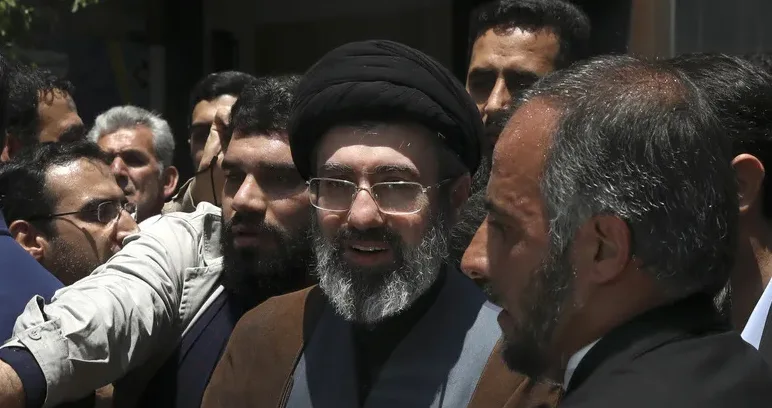 Krupni kadar na muslimanskog vjerskog službenika sa crnim turbanom, naočarima i bradom, okruženog drugim muškarcima.