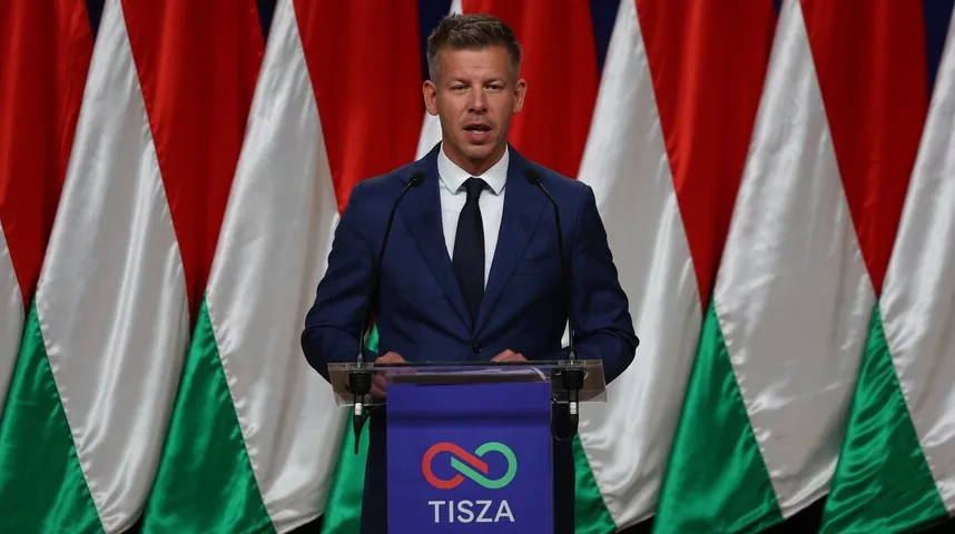 Muškarac u tamnom odijelu sa bijelom košuljom i crnom kravatom stoji za govornicom. Na govornici je ljubičasti dio sa znakom beskonačnosti u crvenoj i zelenoj boji, te natpisom "TISZA". U pozadini se nalaze zastave koje su pretežno crvene, bijele i zelene boje, nalik na zastavu Mađarske.