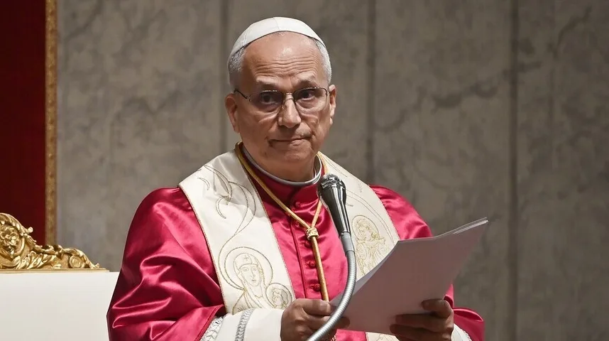 Nadbiskup u crvenoj halji i bijelom pojasu sa zlatnim ukrasima, sa naočalama i bijelom kapom, drži papir u ruci dok govori iza mikrofona.