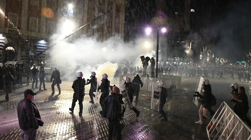 Policija u punoj opremi, sa šljemovima i štitovima, koristi suzavac tokom nemira na ulici, dok pada kiša. U pozadini se vidi veliki broj policajaca i okupljenih ljudi.