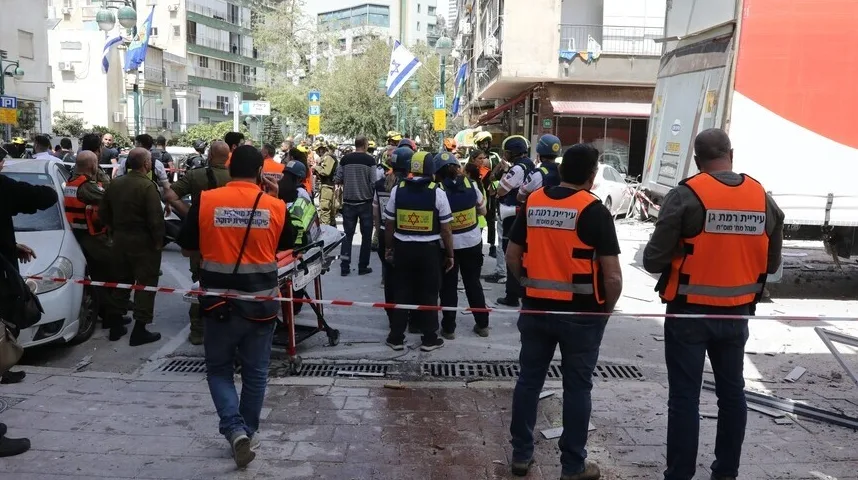 Grupa ljudi u narandžastim prslucima sa oznakama hitne pomoći i vatrogasaca stoje na ulici u blizini srušenog kamiona.